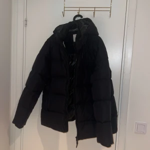 Svart dunjacka från GANT - Säljer en svart dunjacka från GANT, perfekt för kalla dagar. Jackan har en dragkedja framtill och en stilren design med en diskret logga på ärmen. Den är vadderad för extra värme och komfort.