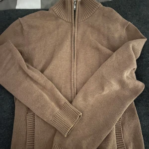 Brun stickad kofta från Dressmann - Säljer en brun stickad kofta från Dressmann med dragkedja och ribbade detaljer vid krage och ärmslut. Perfekt för kyligare dagar. Koftan har en klassisk design som passar till många tillfällen.