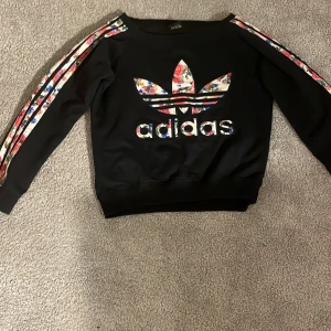 Svart tröja från Adidas med blommigt mönster - Säljer en svart tröja från Adidas med ett stort blommigt logotyptryck på framsidan. Tröjan har långa ärmar med blommiga ränder längs sidorna. Perfekt för en sportig och trendig look!Tröjan är lit offshoulder(stav).Storlek S men passar XS🩷 Inget tecken på användning