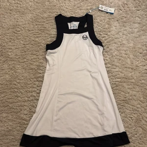 Vit ärmlös tennis klänning från Sergio Tacchini - Snygg vit tennis klänning från Sergio Tacchini med svarta detaljer. Perfekt för sportiga tillfällen med sin ärmlösa design och bekväma passform. Märkets logga pryder framsidan för en stilren look.