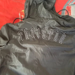 Svart vindjacka från Trapstar - Snygg svart vindjacka från Trapstar med huva och dragkedja. Jackan har en cool broderad logga på ryggen och en rund emblem på bröstet. Perfekt för en streetwear-look.