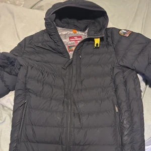 Svart dunjacka från Parajumpers - Säljer en svart dunjacka från Parajumpers med huva och dragkedja. Jackan har en broderad patch på ärmen och är quiltad för extra värme. Perfekt för kyliga dagar.