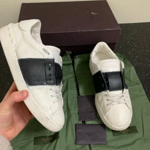 Valentino Open  - Snygga Valentino Open sneakers i vitt med en bred svart rem över ovansidan. Skorna har en klassisk design med snörning och en platt sula. Perfekta för en stilren look. Den är bra skick. Allt og ingår med. Den har allt som kom med från början. Pris är inte hugget i sten. Vid funderingar så är det bara att skriva 