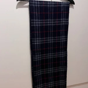 Rutig halsduk från Burberry - Snygg och elegant rutig halsduk från Burberry i mörkblått med inslag av rött och grått. Tillverkad i 100% kashmir för en lyxig och mjuk känsla. Perfekt för att hålla dig varm och stilren under kyliga dagar. Fransen ger en extra touch av elegans.