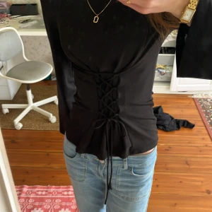 Svart topp med snörning från Gina Tricot - Snygg svart topp från Gina Tricot med snörning framtill som ger en cool look. Toppen har långa ärmar och en figurnära passform. Perfekt för att styla med jeans eller kjol för en trendig outfit.