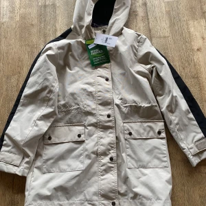 Beige och svart regnjacka med huva - Säljer en beige och svart regnjacka med huva från Cubus. Jackan har knappar framtill och stora fickor med lock. Den är designad för att vara vattenavvisande. Perfekt för regniga dagar!