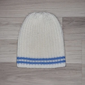 Vit stickad mössa med blå ränder - Säljer en vit stickad mössa med två blå ränder längst ner. Perfekt för kyliga dagar och ger en stilren look. Mössan har en ribbad struktur som ger extra värme och komfort. Hemmastickad.
