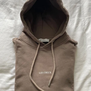 Brun hoodie från Les Deux - Säljer en stilren brun hoodie från Les Deux med justerbar huva och snören. Perfekt för en avslappnad look. Den har en enkel design med märkesloggan tryckt på bröstet. Använd 2-3 gånger, så i nyskick. 