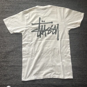 Stüssy t shirt - Säljer en vit t-shirt från Stüssy med deras ikoniska logga tryckt både fram och bak. T-shirten är kortärmad och tillverkad i bomull, perfekt för en avslappnad stil. Liten fläck vid mitten av tröjan men kan enkelt tvättas bort. 