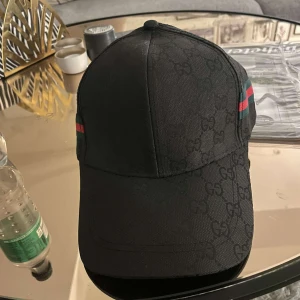 Svart keps med mönster och ränder - Snygg svart keps med diskret mönster och ikoniska ränder i grönt och rött på sidan. Perfekt för en stilren look.