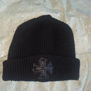 Chrome Hearts Beanie - Snygg svart ribbad mössa med en broderad korsdetalj framtill och en metallknapp på sidan. Perfekt för att hålla värmen med stil.
