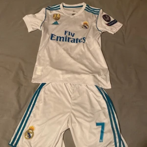 Real Madrid fotbollströja med nummer 7 - Säljer en vit Real Madrid fotbollströja från Adidas med korta ärmar. Tröjan har blå detaljer och nummer 7 på ryggen. Den har även klubbens emblem och sponsorlogga på framsidan samt Champions League-märke på ärmen. Inga skador eller liknande 