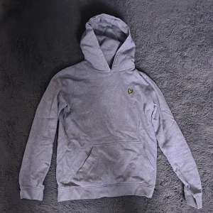 Grå hoodie från Lyle & Scott - Säljer en stilren grå hoodie från Lyle & Scott med deras ikoniska gula logga på bröstet. Tröjan har en klassisk känguruficka och en bekväm huva. Perfekt för en avslappnad stil.