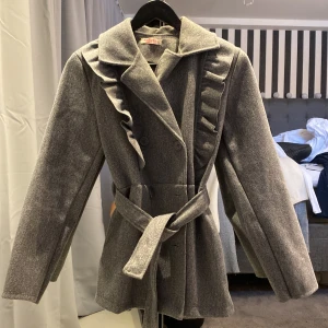 Grå kappa - Elegant grå kappa med volangdetaljer längs kragen och framsidan. Kappan har ett bälte i midjan för en snygg passform och knappar framtill. Perfekt för kyligare dagar.