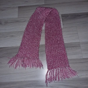 Rosa stickad halsduk - Säljer en mysig rosa stickad halsduk med fransar i ändarna. Perfekt för att hålla sig varm under kyliga dagar. Halsduken har en härlig textur och en fin blandning av rosa och vita toner.