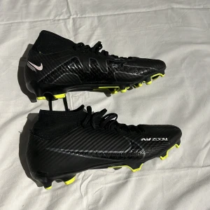 Svarta Nike fotbollsskor med gula dobbar - Snygga svarta Nike fotbollsskor med gula dobbar och snörning. Skorna har en modern design med Nike-logga och texten 'Air Zoom' på sidan. Perfekta för fotbollsplanen med sin greppvänliga sula.