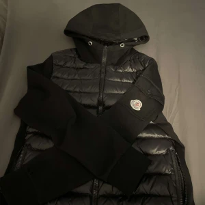 Svart pufferjacka från Moncler - Snygg svart pufferjacka från Moncler med huva och dragkedja. Jackan har långa ärmar och en quiltad design. Perfekt för en vår/sommardag med stil.