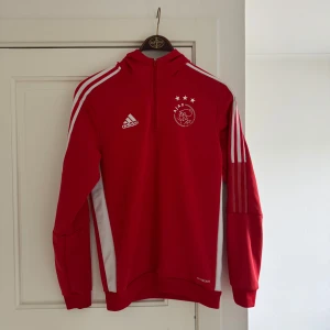 Röd Ajax hoodie - Snygg röd hoodie från Adidas med Ajax-logga på bröstet. Tröjan har en halv dragkedja och vita detaljer på ärmarna. Perfekt för fotbollsfans som vill visa sitt stöd. Den har långa ärmar och en bekväm passform.