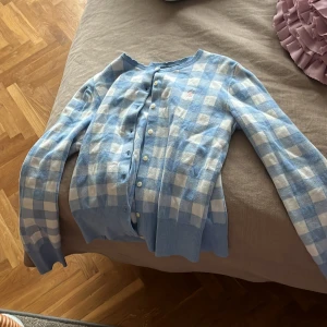 Blå och vit rutig kofta från Ralph Lauren - Säljer en snygg blå och vit rutig kofta från Ralph Lauren. Koftan har långa ärmar och knappar framtill. Den är dekorerad med en liten broderad logotyp i rosa på bröstet. Perfekt för en stilren look!