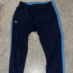 Blå träningsbyxor från Lacoste - Snygga blå träningsbyxor från Lacoste med ljusblå sidopaneler. Perfekta för en aktiv livsstil. Byxorna har en normal passform och är tillverkade i ett bekvämt syntetmaterial.