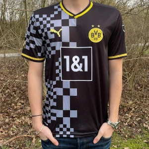 Svart Borussia Dortmund fotbollströja - Snygg svart Borussia Dortmund tröja med gula detaljer och ett coolt rutmönster på ena sidan. Tröjan har korta ärmar och klubbens emblem på bröstet. Nummer 22 och namnet Bellingham pryder ryggen. Perfekt för fans! 🖤💛köpt på marknad i Grekland ej äkthetsbevis