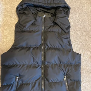 Svart dunväst med huva - Snygg svart dunväst med dragkedja och praktiska fickor. Perfekt för kyligare dagar med sin quiltade design och huva som skyddar mot väder och vind.