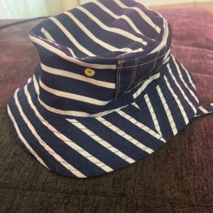 Randig solhatt med gul insida från Chicco  - Snygg solhatt med blå och vita ränder och en överraskande gul insida. Perfekt för soliga dagar med stil. Hatten har en bred kant och dekorativa knappar på sidan.