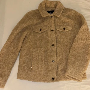 Beige teddyjacka från Hollister - Mysig beige teddyjacka från Hollister med klassisk krage och knäppning framtill. Jackan har två bröstfickor med lock och knappar samt två sidofickor. Perfekt för kyliga dagar och ger en trendig look.