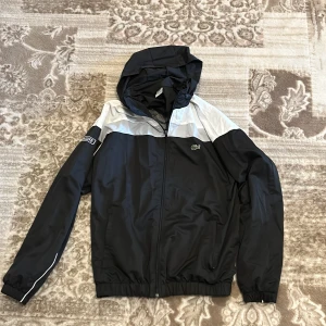 Svart och vit vindjacka från Lacoste - Snygg svart och vit vindjacka från Lacoste med huva och dragkedja framtill. Jackan har logga på bröstet och ärmarna samt elastiska muddar vid ärmslut och midja. Perfekt för blåsiga dagar.