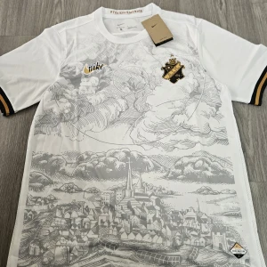 Vit AIK fotbollströja med stadsmotiv - Snygg vit AIK fotbollströja från Nike med svart och guld klubbmärke på bröstet. Framsidan har ett detaljerat stadsmotiv i grått och guldiga detaljer på ärmsluten. Perfekt för dig som vill sticka ut på läktaren eller i stan.