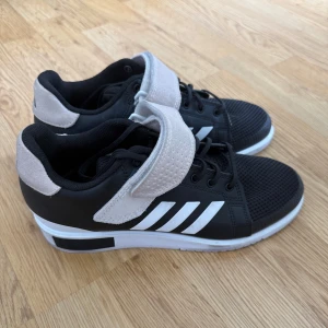 Svarta sneakers med vita detaljer från Adidas - Säljer ett par svarta Adidas sneakers med vita ränder på sidorna och vit sula. Skorna har en bred kardborreknäppning och grå detaljer vid häl och rem. Perfekta för en sportig look.