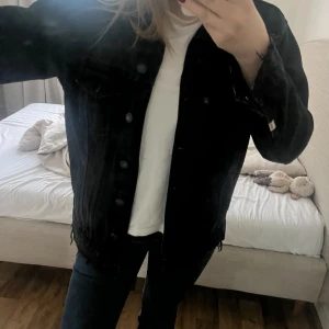 Svart jeansjacka från Bershka - Säljer en svart jeansjacka från Bershka i storlek S. Jackan har klassisk krage, knappar framtill, två bröstfickor med lock och råa, fransiga kanter nedtill. Perfekt för dig som gillar en lite tuffare stil.