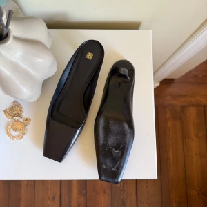 Totême mules med fyrkantig tå - Stilrena svarta mules från Totême. De har låg klack och fyrkantig tå. Skorna har en minimalistisk design och är tillverkade i ett slätt material som ger en elegant look. Perfekta för dig som gillar klassiska och trendiga skor.