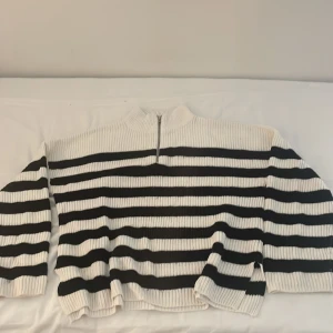 Randig stickad tröja med halv dragkedja - Säljer en vit och svart stickad tröja med breda horisontella ränder och halv dragkedja vid halsen. Tröjan har lång ärm och ribbade muddar. Perfekt för dig som gillar en klassisk och avslappnad stil.