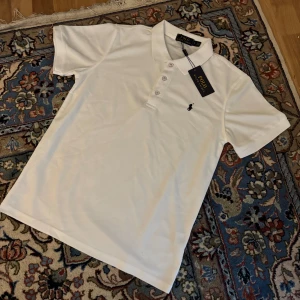 Vit piké från Polo Ralph Lauren - Klassisk vit pikétröja från Polo Ralph Lauren med broderad logga på bröstet. Tröjan har korta ärmar, krage och knappar framtill. Perfekt för en stilren och tidlös look.
