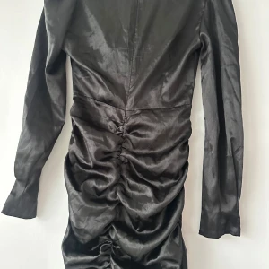 Svart långärmad satin klänning med twist-detalj - Säljer en svart långärmad klänning i satin med twistad detalj framtill och cut-out vid midjan. Klänningen har rynkade partier längs kjolen och en figurnära passform. Perfekt för dig som vill sticka ut med en trendig look.