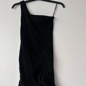Svart one shoulder klänning - Säljer en svart one shoulder klänning med smal axelrem och draperad effekt längs framsidan. Klänningen har en tight passform och är sydd i ett mjukt, lätt ribbat material. Perfekt för dig som vill sticka ut med en stilren look.