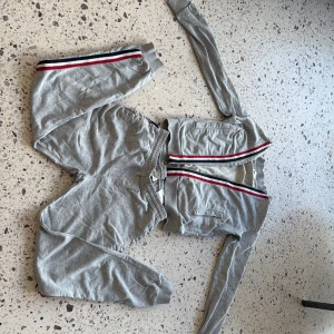 Grå sweatshirt och byxor med ränder från Moncler - Grå långärmad sweatshirt från Moncler med röda, vita och svarta ränder längs ärmar och byxben. Och klassisk Moncler-logga på armen. Perfekt för en sportig och avslappnad stil. (Byxorna och koftan kan också säljas separat)