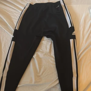 Svarta Adidas träningsbyxor med dragkedja - Svarta träningsbyxor från Adidas med vita ränder längs sidorna och logga på låret. Byxorna har dragkedjor längs bensluten och är tillverkade i ett syntetmaterial som passar perfekt för träning. Klassisk sportig look med smal passform. Sizen är medium 