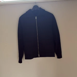 Svart cardigan  - Säljer en svart hoodie med huva och sned dragkedja framtill. Jackan har lång ärm och enkel, stilren design. Perfekt för dig som gillar en clean look och vill ha något bekvämt till vardags.