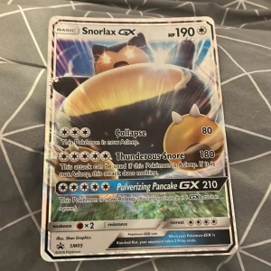 Snorlax GX Pokémonkort - Snorlax GX är ett samlarkort från Pokémon-serien med 190 HP. Kortet har attacker som Collapse, Thunderous Snore och Pulverizing Pancake GX. Framsidan har en bild på Snorlax och kortet har holografiska detaljer.