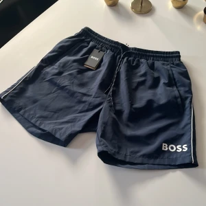 Mörkblå badshorts från BOSS - Snygga mörkblå badshorts från BOSS med vit logga på benet och vita detaljer längs sidorna. Shortsen har elastisk midja med snörning och bakficka. Perfekta för stranden eller poolen. Helt oanvända med lapp!! Kom gärna med offer🙌