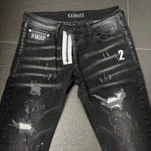 Svarta jeans med slitningar och tryck från XWAY - Säljer ett par svarta jeans från XWAY med slitna detaljer, vita färgstänk och tryck på framsidan. Jeansen har dragkedja och knapp, samt dekorativa detaljer som en vit dragkedja och siffran 2 på ena benet. Snygg streetstil med raka ben.