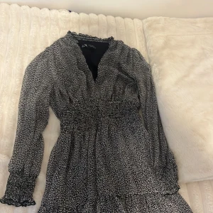 Svartvit mönstrad klänning från Zara - Supersöt svartvit klänning från Zara i storlek XS. Klänningen har långärmade ärmar med smock och volangdetaljer, v-ringad hals och ett småprickigt mönster. Perfekt för dig som gillar feminina detaljer och sköna material.