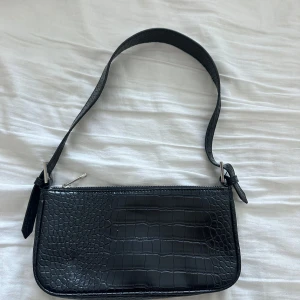 Svart axelväska i krokopräglat mönster från Gina Tricot - Säljer en svart axelväska med krokopräglat mönster. Väskan har dragkedja upptill och ett brett axelband. Perfekt för att bära det viktigaste och ger en stilren touch till din outfit.