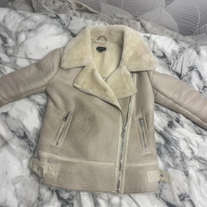 Beige pälsfodrad jacka från New Look - Säljer en beige jacka från New Look med mjukt pälsfoder och stora slag. Jackan har dragkedja framtill, två snedställda fickor med dragkedja och spänndetaljer vid midjan. Perfekt för kyligare dagar.