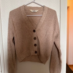 Beige stickad kofta från H&M - Mysig beige stickad kofta från H&M med v-ringning och fyra stora knappar framtill. Koftan har lång ärm och en croppad passform, perfekt att slänga över en topp eller klänning.