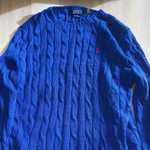 Blå kabelstickad tröja från Polo Ralph Lauren - Säljer en blå kabelstickad tröja från Polo Ralph Lauren i pima cotton. Tröjan har rund halsringning, lång ärm och klassisk röd broderad logga på bröstet. Perfekt för dig som gillar stilrena och tidlösa plagg. Storlek XL