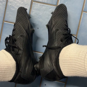 Svarta Nike fotbollsskor med snörning - Säljer ett par svarta Nike fotbollsskor med högt skaft och snörning. Skorna har en modern design med diskret Nike-logga på sidan och mönstrad ovandel för extra grepp på planen. Påse tillkommer 