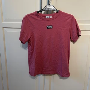 Rosa vintage t-shirt från Adidas - Rosa vintage t-shirt från Adidas med klassisk logga på bröstet och ljusa ränder på ärmen. T-shirten har rund halsringning och är tillverkad i mjukt bomullsmaterial. Perfekt för en sport mm